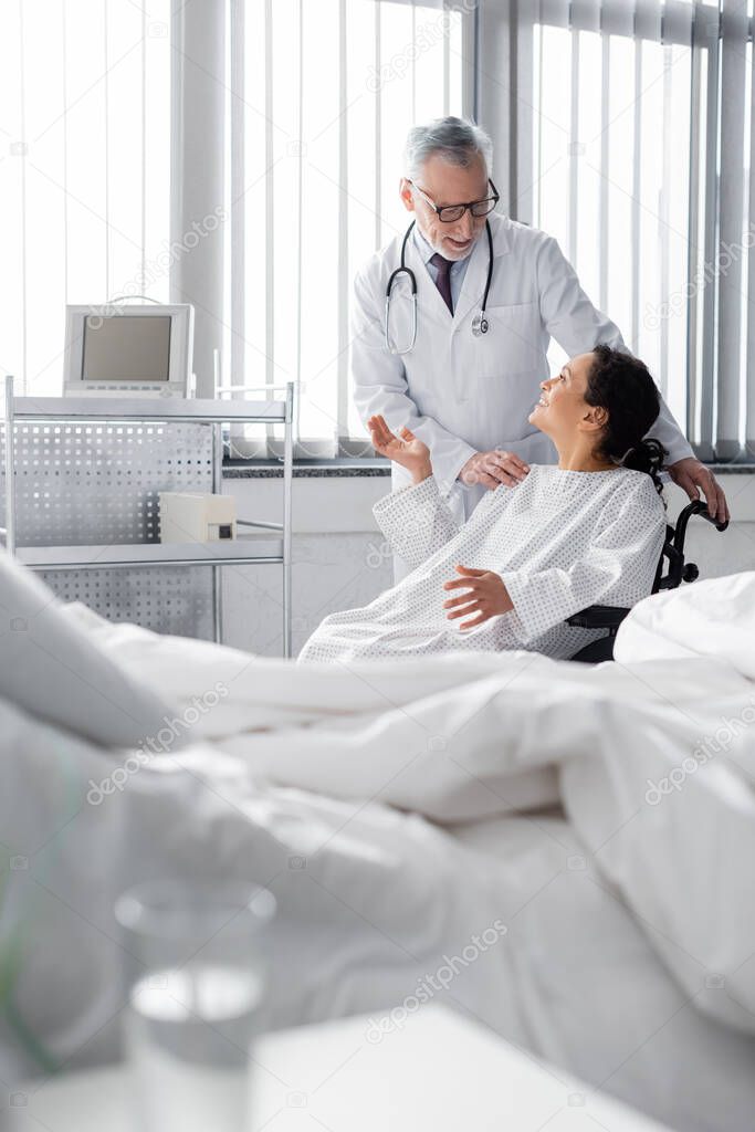 happy african american woman in wheelchair gesturing while talking to doctor near bed on blurred foreground