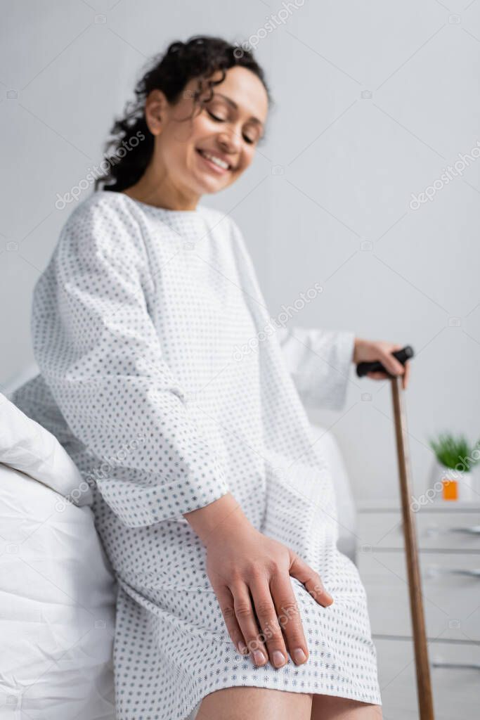 smiling african american woman sitting with walking stick on hospital bed and touching knee, blurred background