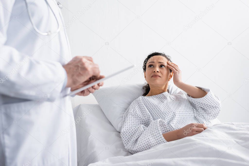 diseased african american woman touching head near doctor with digital tablet on blurred foreground