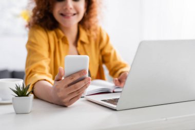 Bulanık arka planda dizüstü bilgisayarın başında çalışırken cep telefonuyla gülümseyen serbest yazar görüntüsü.