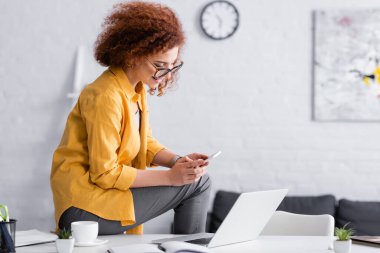 Sırıtan serbest çalışan biri dizüstü bilgisayarın yanında otururken cep telefonuyla sohbet ediyor.