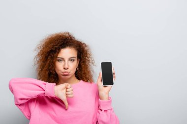 Gri ekranda boş ekranlı akıllı telefonu tutarken baş parmağını aşağı gösteren rahatsız bir kadın
