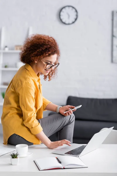 Sonriente Freelancer Escribiendo Portátil Mientras Está Sentado Escritorio Con Teléfono — Foto de Stock