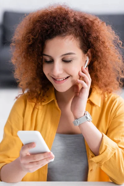 Curly Smiling Freelancer Adjusting Earphone While Holding Mobile Phone — Stock Photo, Image