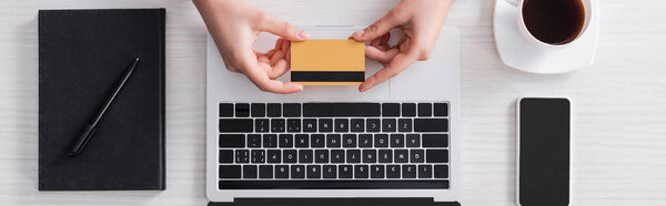 cropped view of freelancer holding credit card near smartphone, laptop and notebook, banner