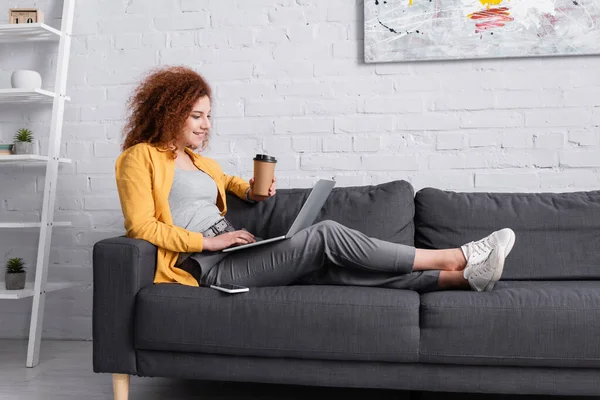卷曲的女人坐在沙发上 带着笔记本电脑和咖啡去 — 图库照片