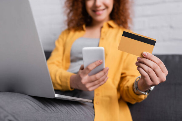 partial view of woman holding smartphone and credit card near laptop on blurred background