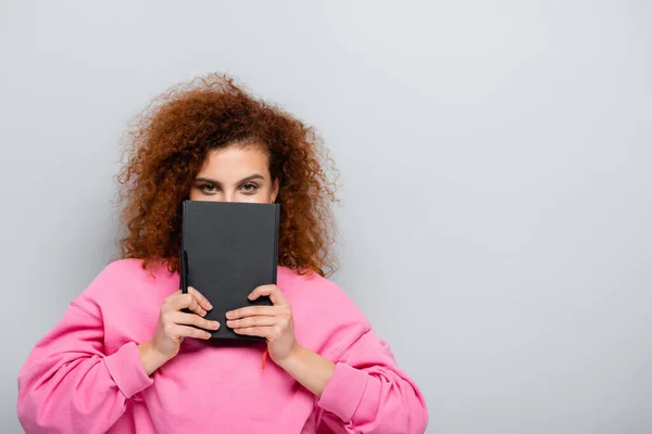 Curly Woman Obscuring Face Notebook While Looking Camera Isolated Grey — Stock Photo, Image