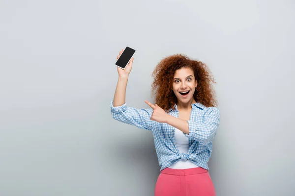 Zdumiona Kobieta Wskazująca Palcem Trzymając Smartfona Pustym Ekranem Szarym Tle — Zdjęcie stockowe