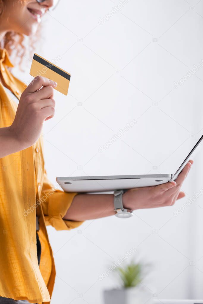 partial view of smiling woman holding credit card and laptop, blurred background