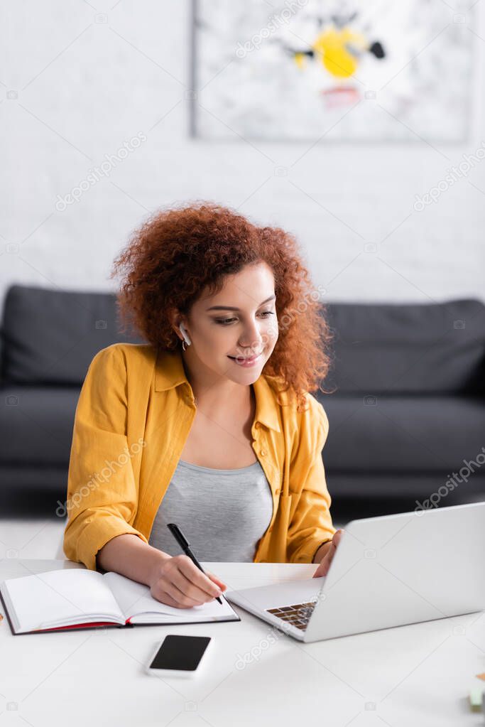 freelancer with earphone writing in notebook near laptop at home