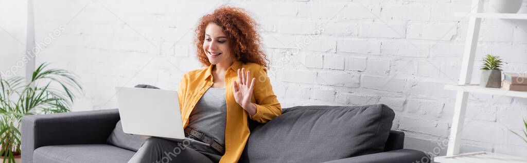 joyful woman having video chat on laptop and waving hand on couch, banner