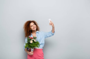 Buketli mutlu kadın gri arka planda akıllı telefondan selfie çekiyor.