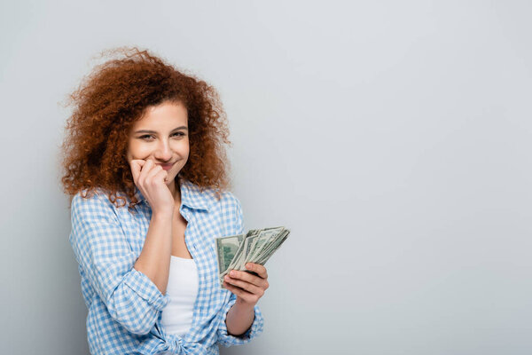 радостная женщина с рукой рядом с лицом держа доллары на сером фоне