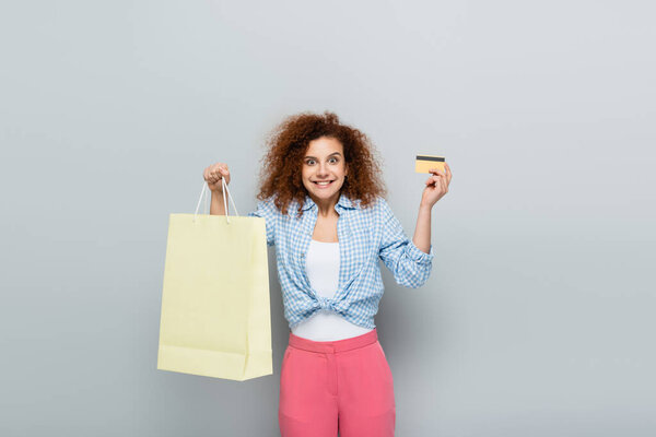 excited woman with curly hair holding credit card and shopping bag on grey background