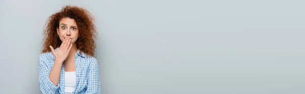 Заплутана Жінка Хвилястим Волоссям Покриває Рот Рукою Ізольовано Сірому Банер — стокове фото