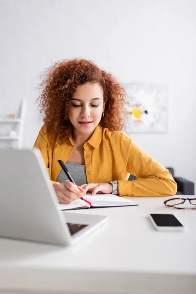 Joven Freelancer Escribiendo Notebook Cerca Smartphone Laptop Primer Plano Borroso —  Fotos de Stock