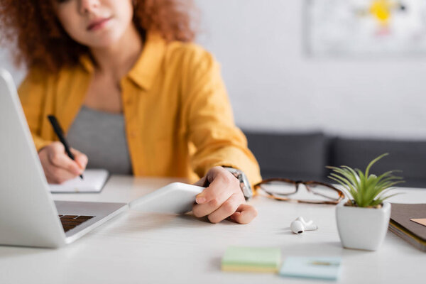 cropped view of freelancer with smartphone working near laptop on blurred background