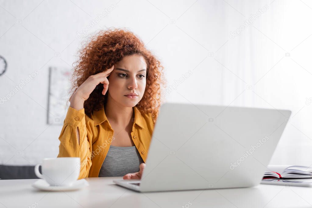 serious freelancer touching head while thinking near laptop on blurred foreground