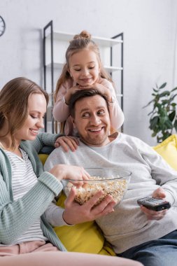 Elinde patlamış mısır ve uzaktan kumandayla gülümseyen adam karısı ve çocuğunun yanında oturuyor. 