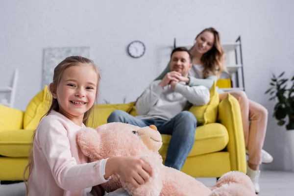 Smiling Kid Holding Teddy Bear Parents Blurred Background — Stock Photo, Image