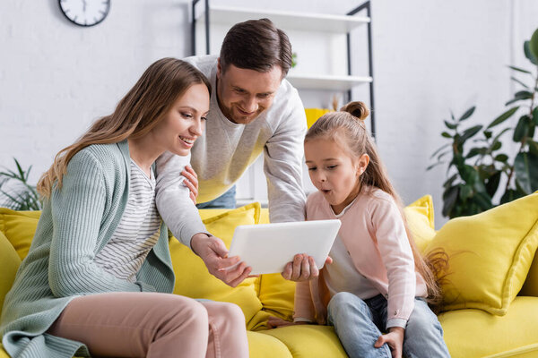 Cheerful woman and daughter looking at digital tablet near husband in living room 