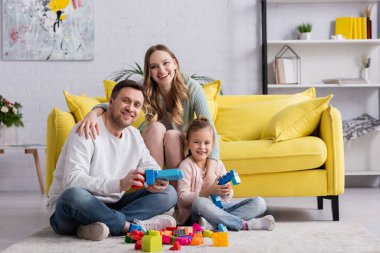 Aile oturma odasındaki yapı taşlarıyla oynarken kameraya gülümsüyor 