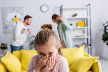 Scared child standing near parents quarrelling in living room on blurred background  clipart