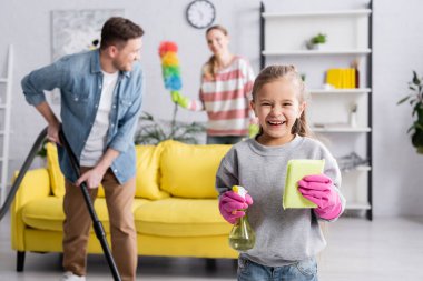 Plastik eldivenli gülümseyen çocuk arka planda ailesinin yanında deterjan ve paçavra tutuyor. 
