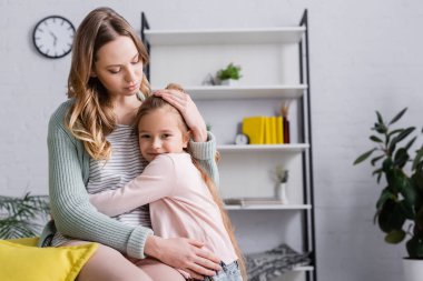 Çocuk evde annesine sarılırken kameraya bakıyor. 