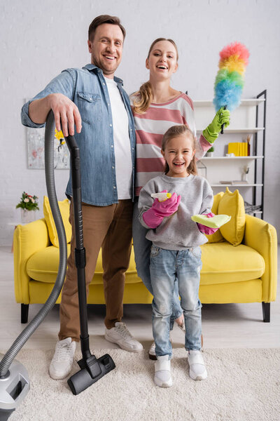 Happy family with detergent, vacuum cleaner and dust brush looking at camera 