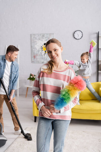 Smiling woman holding dust brush near family on blurred background 