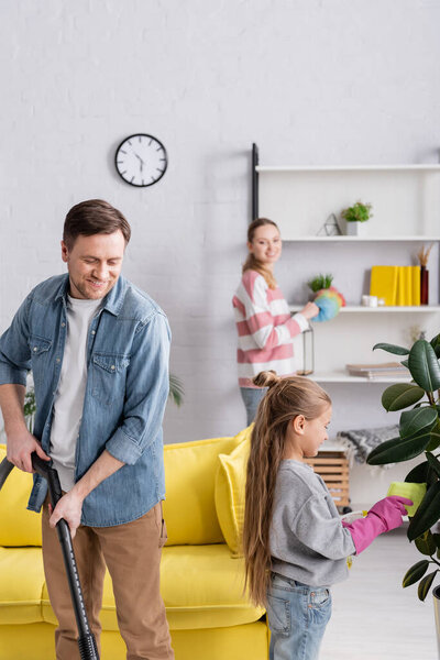 Smiling man using vacuum cleaner near daughter cleaning plant 