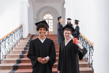 Positive interracial students holding diplomas in university  clipart