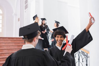 Neşeli Afro-Amerikan mezunu bulanık ön planda arkadaşının yanında diploma tutuyor. 