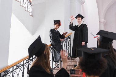 Diplomalarıyla gülümseyen çok ırklı mezunlar bulanık önplanda arkadaşlarına el sallıyorlar. 