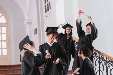 Excited graduate pointing at diploma near interracial friends  clipart