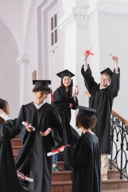 Bulanık ön planda, çok kültürlü arkadaşlarının yanında diplomayı işaret eden neşeli öğrenci. 