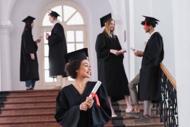 Gülümseyen Afro-Amerikan mezunu. Geçmişi bulanık arkadaşlarının yanında duran diploması var. 