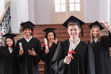 Kep takmış gülümseyen öğrenci arka planda ırklar arası dostların yanında diploma tutuyor. 