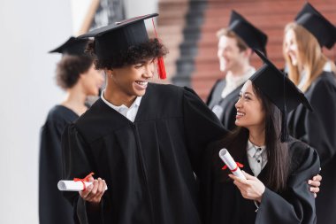 Afrikalı Amerikalı mezun Asyalı arkadaşına diplomasıyla sarılıyor. 