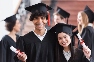 Asyalı arkadaşının yanında gülümseyen diplomalı Afrikalı Amerikalı bekar.  