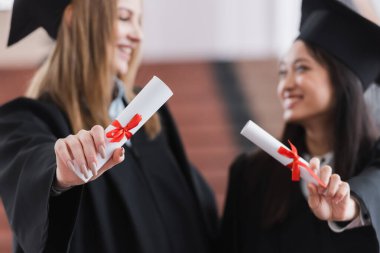 Diplomas in hands of multicultural graduates on blurred background  clipart