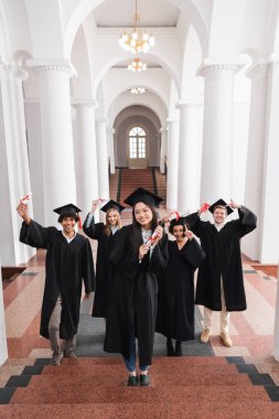 Üniversitede arkadaşlarının yanında diplomasını elinde tutan gülümseyen Asyalı mezun. 