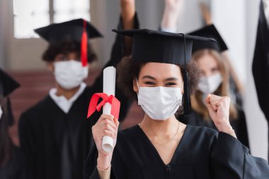 Afro-Amerikan tıp maskesiyle mezun oldu. Elinde diploma var ve evet işareti yapıyor. 