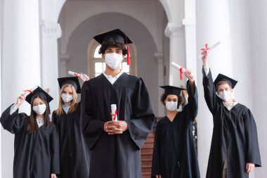 Tıbbi maske takmış Afro-Amerikan mezunu. Geçmişi bulanık arkadaşlarının yanında diploma tutuyor. 