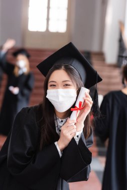 Akademik elbise ve tıp maskesi içinde diplomasını kurdeleyle tutan Asyalı bir mezun. 