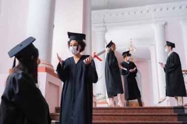 Tıbbi maskeli Afrikalı Amerikalı öğrenci diplomasını tutuyor ve Asyalı arkadaşıyla konuşuyor. 