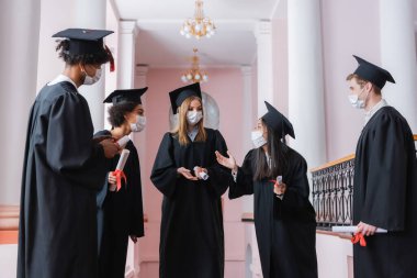 Koruyucu maskeli Asyalı mezun, diplomalı ırklar arası arkadaşlarla konuşuyor. 