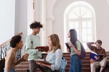 Afrikalı Amerikalı öğrenci Asyalı arkadaşıyla konuşuyor. Not defteri ve akıllı telefonu var. 